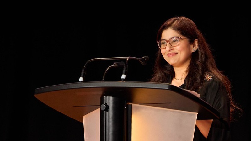 Photograph of Deepa Matoo standing at a podium and smiling.